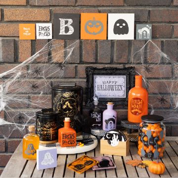 Show details for Spooky Scenes A Halloween-themed display with potion bottles, candy jars, spell books, and framed art on a wooden table, set against a brick wall decorated with cobwebs, spiders, and Halloween signs featuring pumpkins, ghosts, and festive messages.