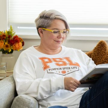 Show details for Pumpkin Spice Life A person with short, gray and purple hair, wearing yellow glasses and a white "PSL Pumpkin Spice Life" sweatshirt, sits on a couch reading a book. A bouquet of orange flowers is on the table beside them.