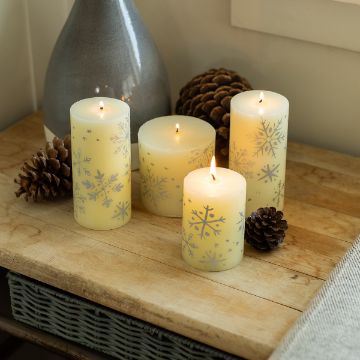 Show details for Special Snowflakes Four white candles with silver snowflake designs are lit and placed on a wooden table alongside pinecones, near a ceramic vase and a window.