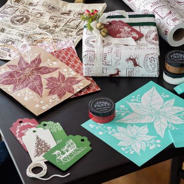 Show details for Etched Christmas Essentials A table displays festive holiday gift-wrapping supplies, including stamped paper, gift tags, kraft bags, paint jars, and a wrapped present decorated with a red poinsettia and green ribbon.