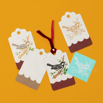 Show details for Bird Minis Four decorative gift tags with bird illustrations and scalloped edges are arranged on an orange background. One tag has a red ribbon, and a turquoise stencil with a bird design is placed nearby.