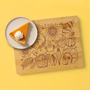 Show details for Fall Favorites A slice of pumpkin pie with whipped cream on a plate sits on a cutting board with engraved fall-themed illustrations, including sunflowers, pumpkins, leaves, croissants, and a basket, on a yellow background.