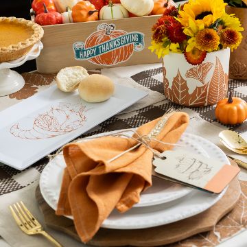 Show details for Thanksgiving Harvest A festive Thanksgiving table setting with orange napkins, gold cutlery, a roll, a pie, a floral arrangement, small pumpkins, and a "Happy Thanksgiving" sign in a wooden box.
