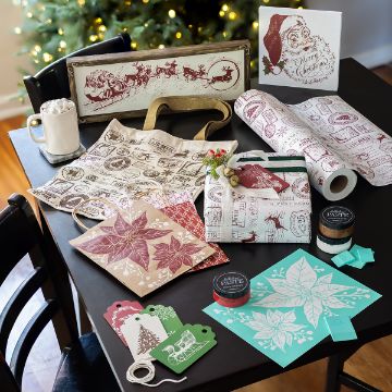 Show details for Poinsettias A table with Christmas-themed wrapping paper, gift bags, tags, stencils, jars of paste, a tote, cocoa with marshmallows, and festive decor. A decorated Christmas tree is visible in the background.