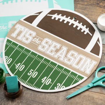 Show details for Football Season Round sign designed like a football and field, with "TIS the SEASON" written across the middle, surrounded by craft supplies including green paint and scissors on a wooden surface.