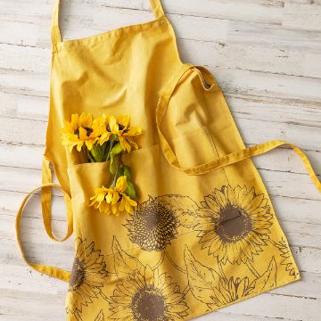 Show details for Sunflower Pattern A yellow apron with sunflower designs lies on a white wooden surface, with several artificial yellow sunflowers placed inside one of its front pockets.