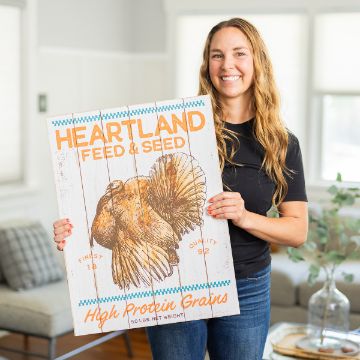 Show details for Heartland Feed & Seed A woman with long wavy hair, wearing a black shirt and jeans, stands indoors holding a rustic wooden sign that reads "Heartland Feed & Seed" and features an illustration of a chicken.