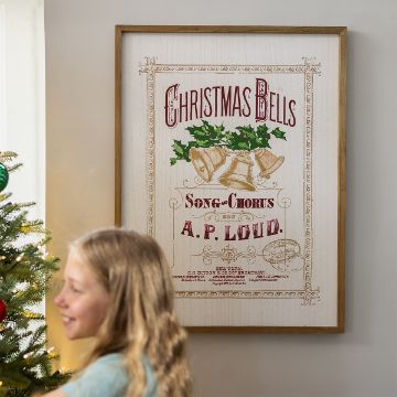 Show details for Christmas Bells A girl stands near a decorated Christmas tree, smiling. On the wall behind her is a framed vintage poster with holly and bells that reads, "Christmas Bells, Song & Chorus by A.P. Loud.