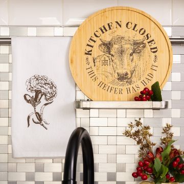 Show details for Kitchen Closed A kitchen scene with a round wooden sign reading "Kitchen Closed, This Heifer Has Had It" featuring a cow illustration, a towel with a flower drawing, and a shelf holding berries and greenery above a sink.