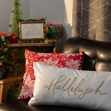 Show details for Hallelujah Script A festive living room scene with a black leather chair, red and white holiday pillows, a small Christmas tree, wrapped gifts, and a framed drawing of the nativity. One pillow reads "Hallelujah" in gold script.