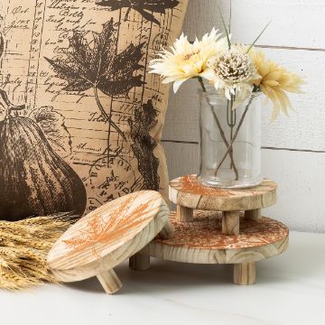 Show details for Riser (3-Pack) A small pillow with botanical prints, a glass jar with yellow flowers, and three round wooden risers with orange leaf designs, all arranged on a white surface with wheat stalks nearby.
