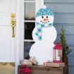 A snowman decoration with a blue snowflake hat and checkered scarf stands by a front door, next to a small pine tree, red lanterns, and wrapped gifts on a wooden box.