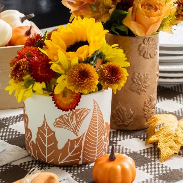 Show details for Leaf Artisan Add-Ons A fall-themed table display featuring a white and brown leaf-patterned vase with sunflowers and chrysanthemums, a tall vase with orange roses, a mini pumpkin, and a checkered tablecloth.