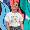 A smiling woman in a white t-shirt that reads "This Girl Loves Jesus" stands in front of a colorful, abstract mural. She has light brown hair tied back and is wearing black-and-white striped pants.