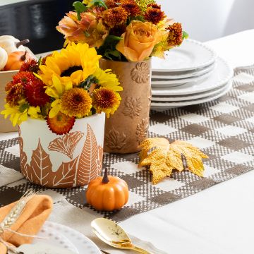 Show details for Modern Plaid Pattern A fall-themed table setting with white plates, gold flatware, orange and yellow flowers in decorative vases, a small pumpkin, and yellow faux leaves on a brown and white checkered table runner.