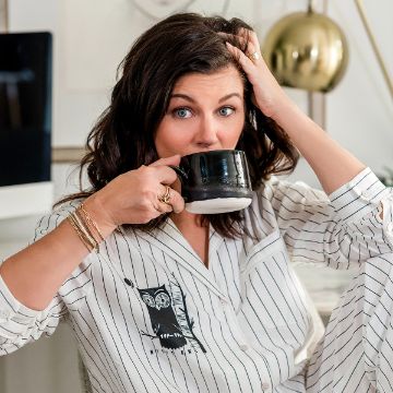 Show details for Night Owl A woman with dark hair wearing striped pajamas with an owl print sips from a black mug, looking surprised. She sits indoors with one hand on her head and the other holding the cup. A gold lamp is in the background.