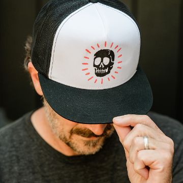 Show details for Caught In Your Web A man with facial hair and a wedding ring tilts his black-and-white cap, which features a cartoon skull with red rays around it. His eyes are not visible as the brim of the hat hides the top half of his face.