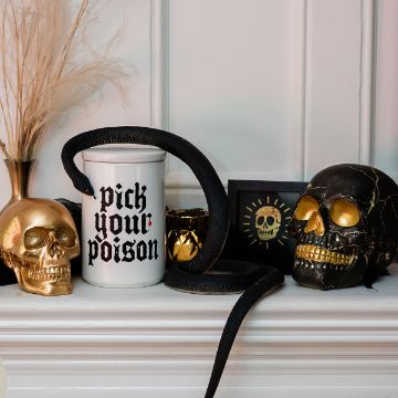 Show details for Pick Your Poison A black snake coils around a white mug labeled "pick your poison," next to gold and black skulls, a gold cup, and framed art with a sun and eye, all arranged on a white mantel.