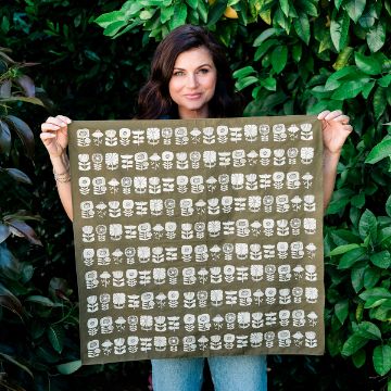 Show details for Sunflower Power A woman stands outdoors in front of leafy green bushes, holding up a square olive-green cloth decorated with rows of white abstract floral and geometric patterns.