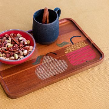 Show details for Fallen Fruit A wooden tray with drawings of a pear and an apple holds a small bowl of mixed nuts and a dark mug with a cinnamon stick, set on a mustard yellow textured surface.