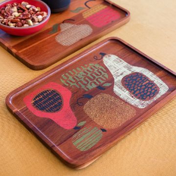 Show details for Feeling Gourd A wooden tray with colorful, stylized fruit designs sits on a yellow surface. Another tray with similar artwork and a bowl of mixed nuts are partly visible in the background.