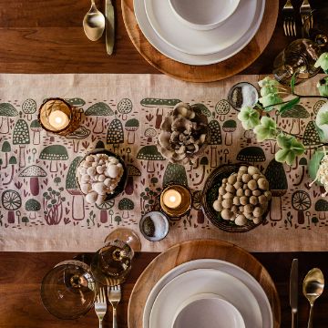 Show details for Magic Mushrooms A wooden dining table set with white plates, gold cutlery, candles, and glasses. The table runner features whimsical mushroom illustrations, and decorative mushrooms and succulents are arranged as a centerpiece.