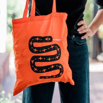 Show details for Snake Bite A person stands with one hand on their hip, holding an orange tote bag featuring a black snake design with stars and a small pink tongue.