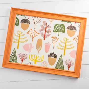 Show details for Falling For You Framed artwork with orange frame featuring a playful pattern of trees, acorns, and abstract plants in green, yellow, pink, and brown tones on a white background, set against a white wood wall.