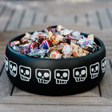 Show details for Bone-afide A black bowl decorated with white skulls is filled with assorted wrapped candies and sits on a wooden table.