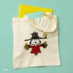 A beige tote bag with an illustration of Snoopy dressed as a scarecrow, wearing a hat and red shirt, is shown on a light green surface with a yellow and a turquoise notebook peeking out from the bag.