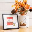 A framed Peanuts illustration with Lucy and Snoopy at a Thanksgiving pie stand sits next to a white vase with Snoopy design, filled with artificial autumn leaves and pinecones, on a wooden surface.