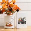A white vase with Snoopy jumping into autumn leaves sits next to a canvas featuring Snoopy, Woodstock, and Charlie Brown with the word "GIVING" in large letters. Fall leaves and pinecones fill the vase.