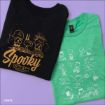 Two folded Peanuts-themed t-shirts on a purple background: one black with gold "Spooky" text and Peanuts characters, and one green with white cartoon sketches of Peanuts characters playing.