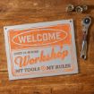 A metal sign on a wooden surface reads: "Welcome. Open 24 hours. Workshop. My tools, my rules." Next to it are a ratchet wrench and three socket attachments.