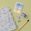 A floral notebook with colored tabs, pastel sticky notes, purple floral washi tape, scattered purple petals, and a gold pen on a light green background.