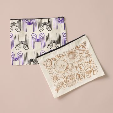 Show details for Zipper Pouch - Club Couture Exclusive Two fabric zipper pouches on a light pink background. One pouch has a black and purple geometric pattern, and the other features brown line drawings of fruits, bread, and baked goods.