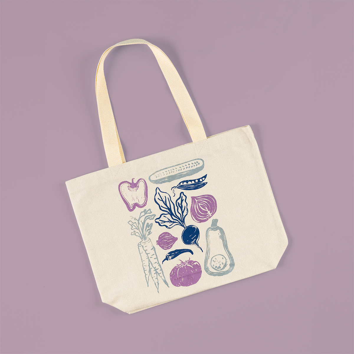 A cream-colored tote bag with illustrations of various vegetables in purple, blue, and green on the front, including a bell pepper, cucumber, pea pod, lettuce, carrot, eggplant, cabbage, tomato, and squash. The background is mauve.