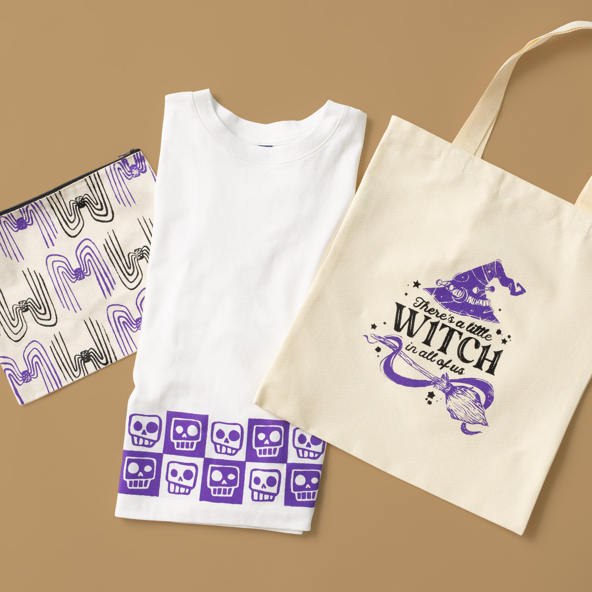 A folded white t-shirt with purple skulls, a cream tote bag with a purple witch hat and text "There's a little witch in all of us," and a patterned pouch with black and purple designs, all on a tan background.