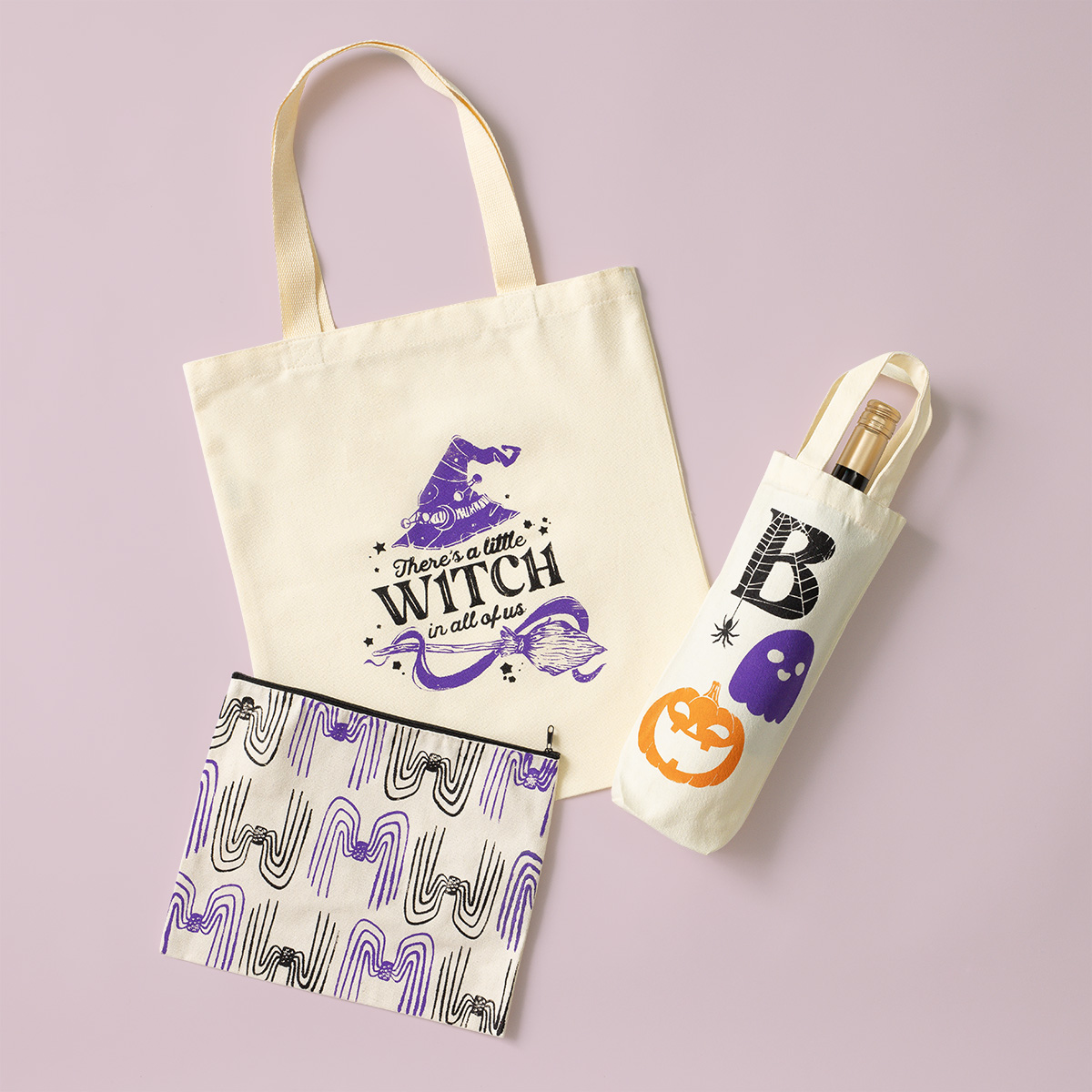 Canvas tote bag with a purple witch hat design and "There's a little witch in all of us" text, a matching pouch with a purple and black pattern, and a wine bag with a ghost, pumpkin, and letter B on a light background.