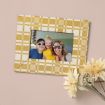 A framed photo of a smiling woman and two children, all wearing sunglasses, is placed on a light pink surface beside a bunch of pale yellow artificial flowers. The frame has a yellow and white checkered pattern.