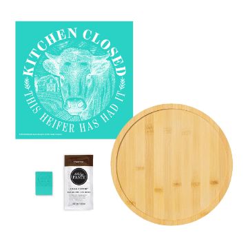 Show details for Kitchen's Closed Try-Me Kit A teal sign reads "Kitchen Closed, This Heifer Has Had It" with a cow illustration, next to a round bamboo cutting board, a small teal square, and a packet labeled "Espresso Cheese Paste.