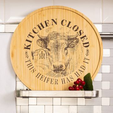 Show details for Kitchen's Closed Try-Me Kit A round wooden cutting board with an engraved cow, barn, and the words “KITCHEN CLOSED, THIS HEIFER HAS HAD IT,” leaning against a white tile backsplash above a metal shelf with berries and green leaves.