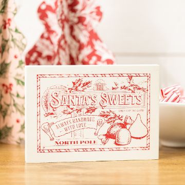 Show details for Santa's Sweets A white box labeled "Santa’s Sweets" with red vintage-style lettering and holiday illustrations sits on a wooden surface, with candy and festive fabric in the background.