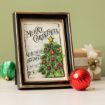 A framed Christmas card featuring a decorated tree and the words "Merry Christmas" stands on a table beside a red ornament, a green ornament, and a wrapped gift with a bell.