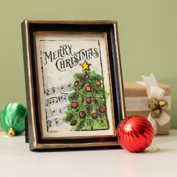 Show details for O Christmas Tree A framed Christmas card featuring a decorated tree and the words "Merry Christmas" stands on a table beside a red ornament, a green ornament, and a wrapped gift with a bell.