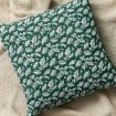 A square pillow with a green cover featuring a white holly and berry pattern, resting on a textured beige knit blanket.