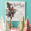 A vintage-style "Joyeux Noël" sign featuring Santa Claus holding gifts is mounted on a turquoise wall above a white table decorated with patterned paper Christmas trees and a bowl of peppermint candies.