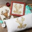 A pillow, a framed print, and a tag display a gold and brown reindeer illustration with holly. Nearby are scissors, paint, ink, and a stencil on a rustic wooden surface.
