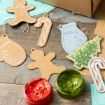 Holiday cookie-shaped ornaments, including mittens, gingerbread people, and candy canes, are laid out on a wooden surface with twine. Open jars of red and green paint are nearby for decorating.