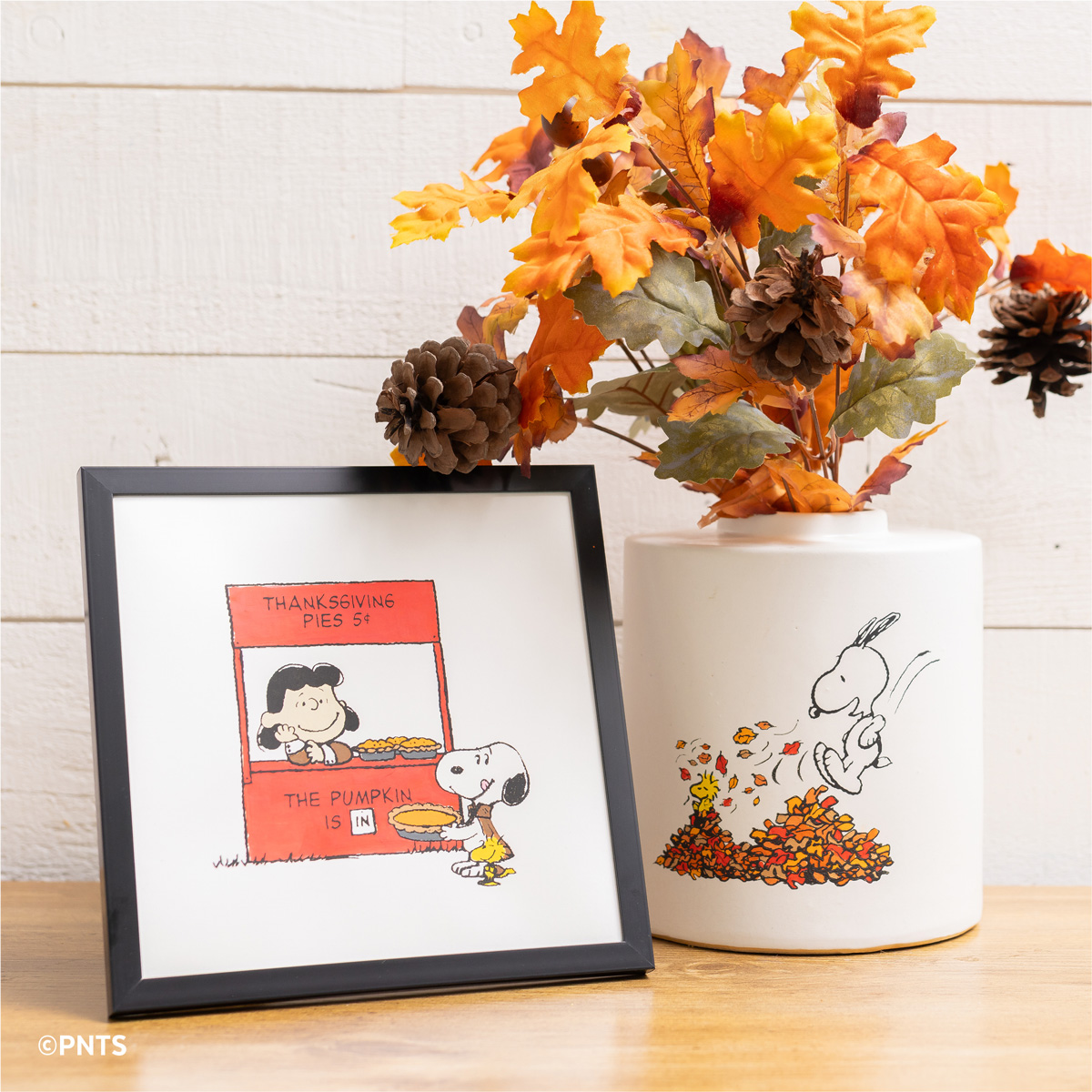A framed Peanuts comic showing Lucy and Snoopy at a pumpkin pie stand sits next to a white vase with Snoopy playing in autumn leaves, filled with fall foliage and pinecones, on a wooden surface.